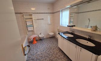 A modern bathroom with two sinks and a large mirror. The walls are light tiled and there is a small window opening for daylight.