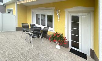 A cozy garden area with a table and chairs in front of a yellow wall. The flowers and the entrance door give the space an inviting atmosphere.