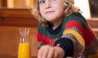 A small boy with curly hair and a yellow cap is sitting at the table. In front of him is a breakfast with rolls, bacon, and a glass of orange juice.