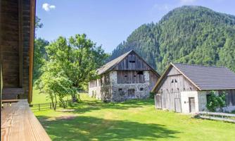 An idyllic farmhouse in a green landscape, surrounded by trees and mountains. The sky is clear and blue, creating a peaceful atmosphere.