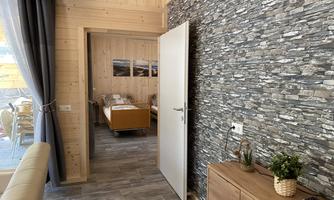 A modern furnished room with a stone wall and wooden elements. In the background, another room with furniture is visible.