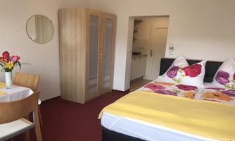 A bright room with a double bed and colorful pillows. A table with flowers and a wardrobe can also be seen.