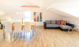 A modern living room with wooden floors and a cozy couch. A dining table with white chairs and colorful cushions creates an inviting atmosphere.