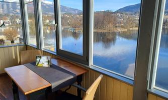 A cozy table with a chair in a bright room. Outside, a calm lake can be seen with mountains in the background.
