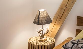 A modern lamp with a shade made of black fabric stands on a table made of wooden logs. The table has a decorative shape with antler applications.