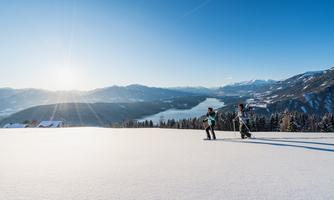 A snowy landscape with a mountain view and a lake in the background. Two people are standing on the slope and enjoying the winter sun.