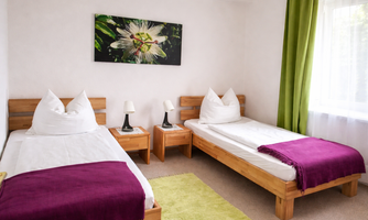 A cozy room with two single beds, furnished with wood. The walls are bright, and a green curtain along with a colorful picture creates a pleasant atmosphere.
