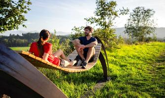 A couple sits relaxed on a modern wooden bench in a green landscape. In the background, trees and gentle hills can be seen.