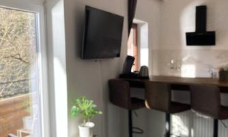 A modern kitchen with bar stools and a TV on the wall. A window lets in natural light and offers a view outside.