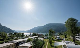 A tranquil landscape with a lake and mountains in the background. RVs and a campsite are visible, under a clear sky with bright sunshine.