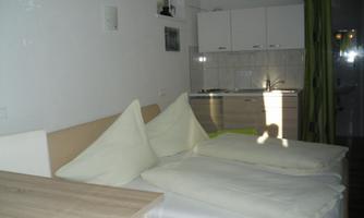 A simple room with a bed and two cushions. The kitchen is visible in the background and the walls are painted light.