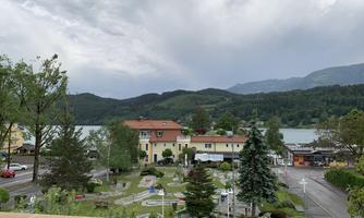 A picturesque landscape with a lake in the background and green mountains. In the foreground, there are a few buildings and well-maintained green areas.