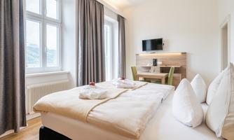 A modern room with a double bed and white bedding. The large window front provides plenty of daylight, and a small workspace is also available.