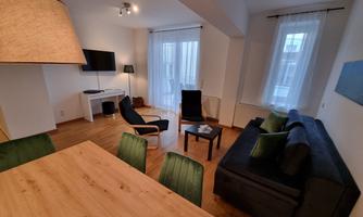 A modern living room with a sofa, a table, and a television. Large windows provide plenty of daylight.
