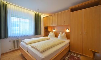 A cozy bedroom with two beds and bright wooden wardrobes. Large windows let in a lot of daylight.