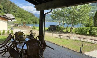 A cozy outdoor area with a table and chairs. In the background, there is a calm lake and green trees visible.
