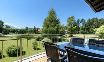 A beautiful balcony with a table and chairs, surrounded by lush greenery. In the background, there are trees and a clear blue sky.