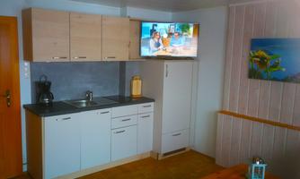 A modern kitchen unit with wooden cabinets and a refrigerator. In the background, a television is visible showing a family eating.