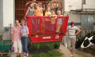 A group of children is standing on a large red trailer in a barn. In the background, cows and agricultural equipment can be seen.