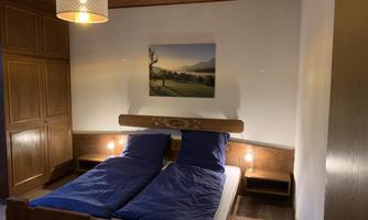 A cozy bedroom with two beds and blue bedspreads. A landscape painting hangs on the wall, and a ceiling lamp provides gentle light.
