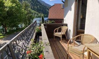A balcony with two chairs and a table, surrounded by plants. In the background, trees and a lake are visible.