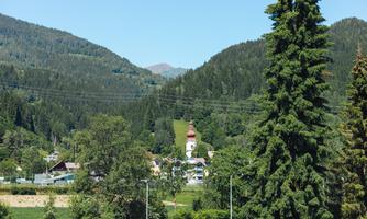 A picturesque mountain landscape with green trees and gentle hills. In the background, a church with a red roof is visible.