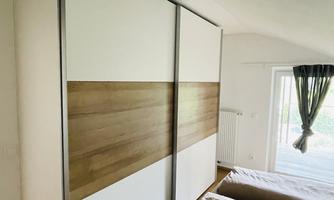 A modern bedroom with a large, elegant wardrobe in white and wood look. In the background, there is a window with curtains that lets in natural light.