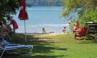 A sunny beach with children playing in the water. In the foreground, there are sun loungers and a beach umbrella.