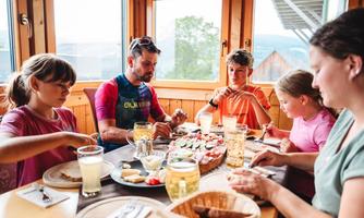 A family is sitting at a table enjoying a hearty meal. They are laughing and eating together while the light streams through the windows.