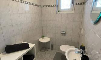 A modern bathroom with white tiles and a large mirror. There is a small shelf with towels and a round table.