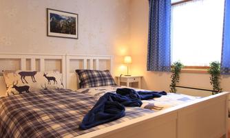 A cozy bedroom with a double bed, plaid bedding, and a warm atmosphere. Large windows let natural light in, and plants add a fresh touch to the room.