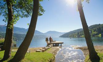 A quiet lake surrounded by trees and mountains. On the shore, two people are standing on a wooden pier.