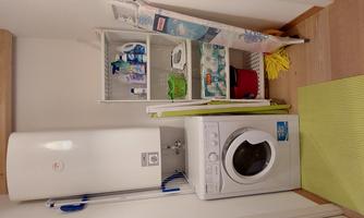 A small laundry room with a washing machine and a sink. Shelves with cleaning products and an ironing board are also available.