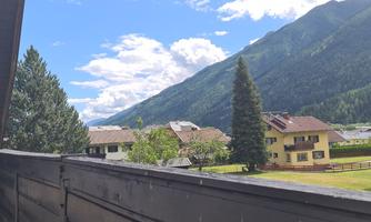 A picturesque mountain landscape with green forests and impressive peaks. In the foreground, some houses and a clear blue sky are visible.