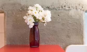 A small vase with white flowers sits on a red table. In the background, a gray wall is visible.