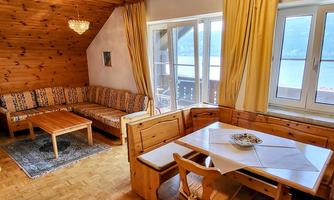 A cozy living room with wooden paneling and large windows. There is a seating area and a dining table with a view of a lake.