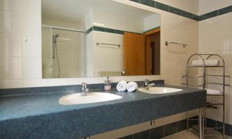 A modern bathroom with two sinks and a large mirror. Towels are neatly arranged, and there is a shower in the background.