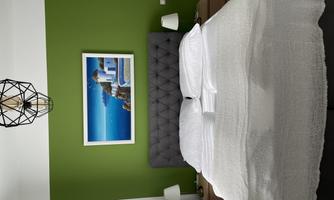 A modern bedroom with a double bed and a gray headboard. The walls are painted green and a picture of a Mediterranean landscape hangs above it.