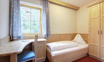 A cozy bedroom with a single bed and a desk. Large windows let in plenty of daylight, and the floor is covered with red carpet.