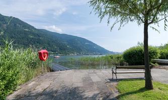 A tranquil lake with green shores and a beautiful mountain horizon. In the foreground, there is a bench and a lifesaver is visible.