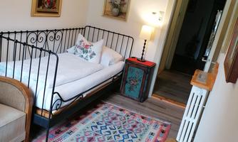 A cozy room with an iron bed and a colorful carpet. Pretty pictures are hanging on the walls, and there is pleasant lighting.