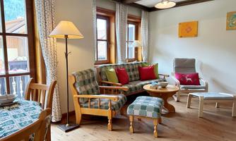 A cozy living room with wooden floors and large windows. The furniture is inviting and colorful, featuring a dining table and a seating area.