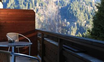 A balcony with a table and a chair, surrounded by an impressive mountain panorama. Nature reveals itself with green forests and a clear sky.