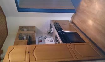 A small kitchen with wood-colored cabinets and a modern cooking area. The floor is made of wood and the walls are in neutral colors.