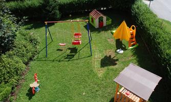 A colorful playground with a swing, a slide park, and a playhouse. Surrounded by green grass and hedges.