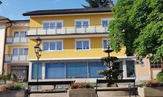 A yellow building with several windows and a balcony. In front of the building, there is a fountain and flowerbeds.