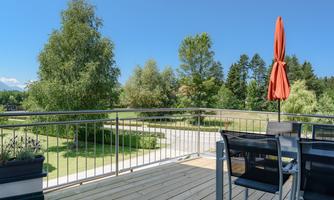 A balcony with modern furniture and a red sunshade. In the background, green trees and a clear blue sky can be seen.