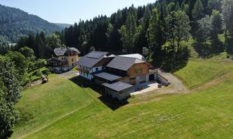A modern house in nature, surrounded by green meadows and trees. In the background, gentle hills and a clear blue sky can be seen.
