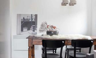 A bright dining room with a wooden table and black chairs. An artwork hangs on the wall, and the lighting consists of modern lamps.