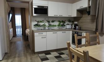 A modern kitchen with white cabinets and a wooden countertop. A wooden dining table stands next to the kitchen, and in the background, a cozy living area can be seen.